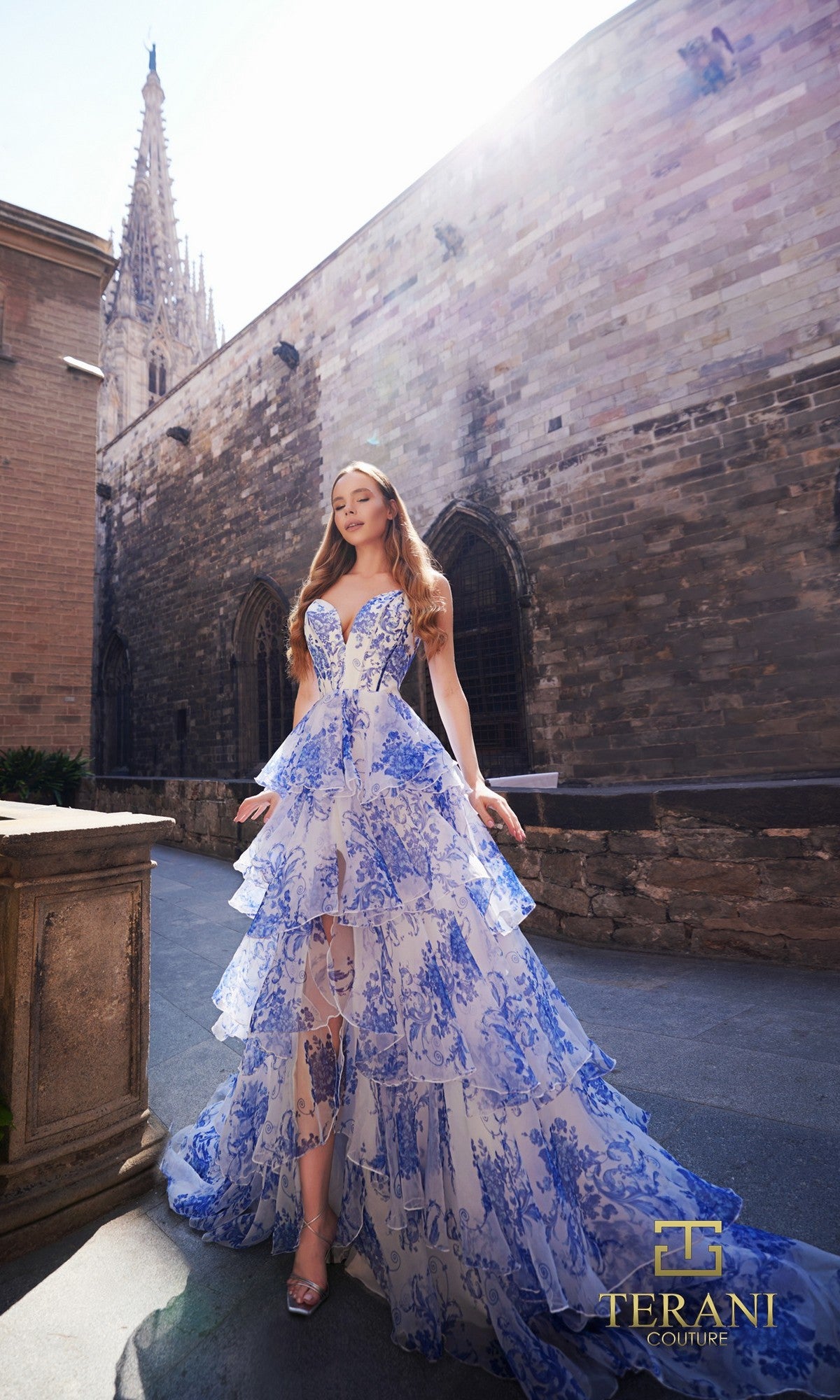 A woman poses outdoors in a blue and white printed chiffon Terani Couture Long Formal Dress 261P6673 with an a-line skirt near a Gothic stone building; the